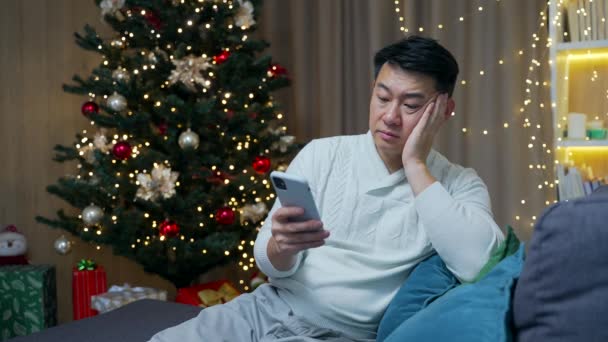 クリスマス休暇に家に座っている退屈アジアの男不幸な疲れた若い男性はXmasを祝うだけで悲しい考えで失われた落ち込んで感情的な孤独感と憂うつな使用スマートフォン電話 — ストック動画