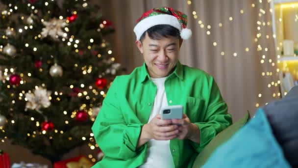Veselý Vtipný Asijský Muž Santa Klobouk Pomocí Smartphone Sedí Doma — Stock video
