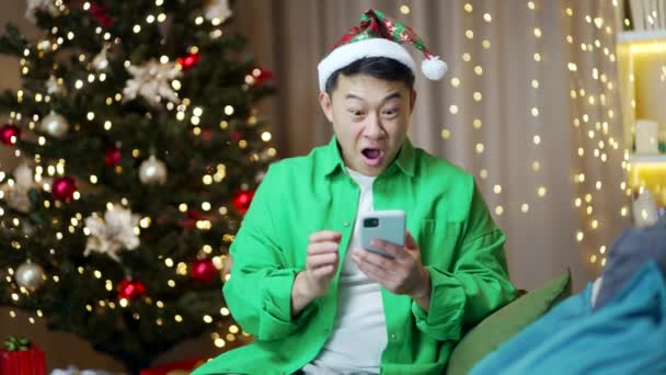 若い興奮したアジアの男幸せな肯定的な笑顔は自宅で座って勝利のスマートフォンXmas冬を祝う喜びます屋内で驚いている携帯電話を見てクリスマスイブ携帯電話で素晴らしいニュースを受け取る — ストック動画