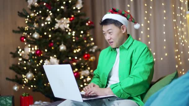 興奮したアジア人は 大規模なオンラインニュースを祝う喜びを感じ ラップトップの画面を見てソファに座って成功を味わい 商業提供を楽しみ 割引を販売し インターネット宝くじの勝利を獲得しました クリスマス — ストック動画