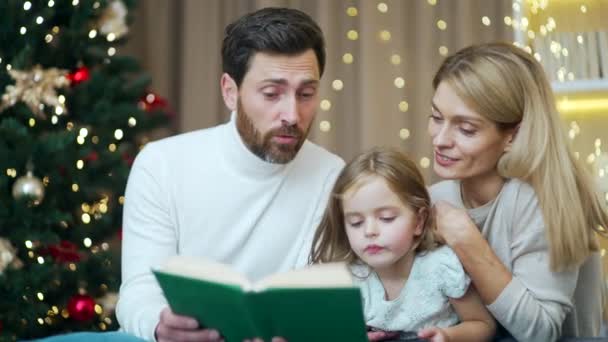 思いやりのある両親はクリスマス イブの居心地の良い家で娘に本を読んでいますソファの背景の新年の木の家に座っています 冬の休日の夜 子供を持つ幸せな家族 — ストック動画