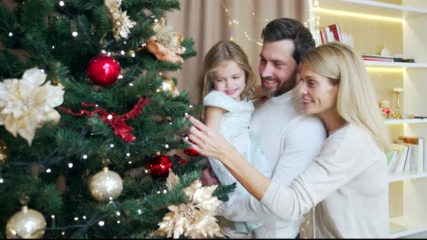 小さな女の子の子供を持つ幸せな若い家族は 居心地の良い家でクリスマスツリーを一緒に飾る 冬の休日クリスマスの伝統と人々の概念 照明を配置し楽しみながら — ストック動画