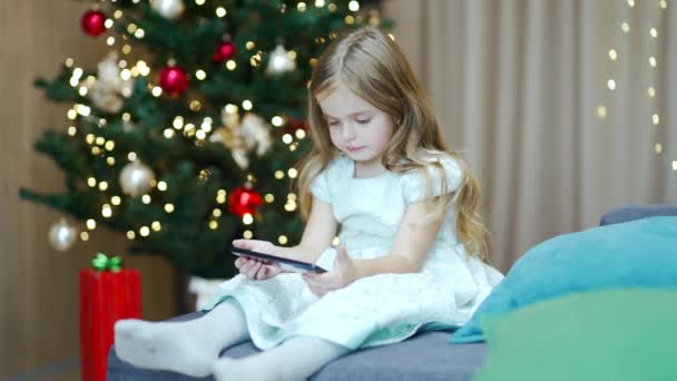 小さな女の子は 自宅の背景クリスマスツリーに座ってスマートフォン上のオンライン興味深いビデオや漫画を見ています かわいい子供が携帯電話を見ている 冬休み中に屋内で子供 — ストック動画