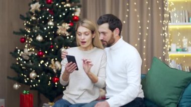 Heyecanlı evli çift mutlu mutlu bir gülümseme kutlayın akıllı telefon xmas kış evde oturup Noel arifesinde cep telefonuna bakmak sürpriz cep telefonu kazanmak kazandı