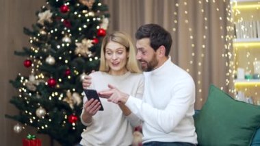 Evli çift Noel hediyelerini online tatil tekliflerinde seçiyor. Mutlu eşler oturma odasındaki parıldayan yeni yıl ağacının yanında oturup aile dostlarına Noel hediyesi almak için bir seçim yaparlar.