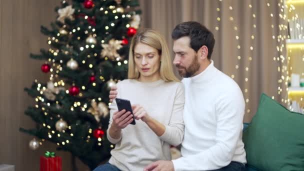 結婚したカップルは休日のオンライン提供のXmasのギフトを選ぶ 幸せな愛情のある配偶者は リビングルームで輝く新年の木に座って電話注文で選択をするクリスマスプレゼント家族の友人のための — ストック動画