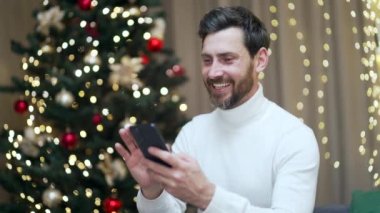 Yakışıklı adam yılbaşı tatilinde evinde koltukta oturup Noel ağacına karşı akıllı telefon kullanıyor. İş adamı, cep telefonu takılıyor, sohbet ediyor, daktilo kullanıyor.