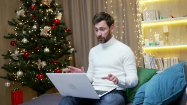 Mutlu ve yakışıklı erkek çalışan serbest çalışan işadamı, yeni yıl tatilinde evde oturan dizüstü bilgisayarla web kamerası görüntüleriyle uzaktan konuşuyor. Konuşur