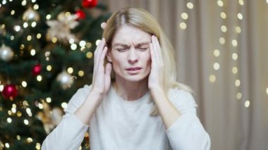 Oturma odasında otururken baş ağrısı çeken bir kadın Noel ağacının arkasında oturuyor. Yılbaşı bayramlarındaki ağrı gerginlik baş ağrısı stresi. Kapalı