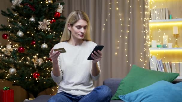 支払いの難しさ オンラインストアで購入しようとしている不満女性 女性はクリスマスにだまされました 自宅でスマートフォンの携帯電話やクレジットカードでソファに座って — ストック動画