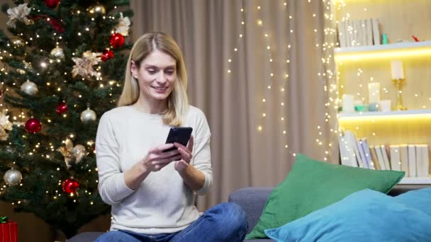 若い興奮した女性幸せな肯定的な笑顔は自宅で座って勝利スマートフォンXmas冬を祝う喜びクリスマス休暇中の携帯電話を見て驚いた携帯電話で素晴らしいニュースを受け取る — ストック動画
