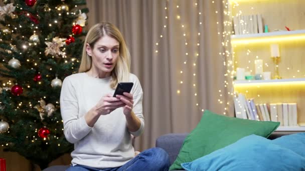 興奮した女性幸せな肯定的な笑顔は自宅で座って勝利のスマートフォンXmas冬を祝う喜びます屋内クリスマス休暇中の携帯電話を見て驚いた携帯電話で素晴らしいニュースを受け取る — ストック動画