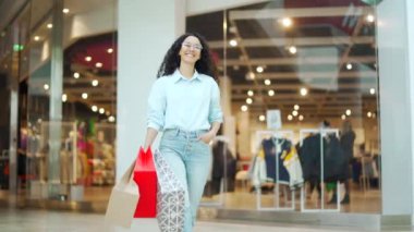 Alışveriş merkezinin önünde duran, alışveriş torbaları taşıyan ve kameraya gülümseyen neşeli genç bir kadının portresi. Kara Cuma 'da alışveriş yapan mutlu bayan neşe içinde atlıyor.