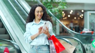 Bir kadın alışveriş merkezinde akıllı telefon kullanıyor. Alışveriş merkezinde alışveriş yapan kadın portresi. Alışveriş torbaları ve uygulamalı akıllı cep telefonu kullanıyor. Mutlu kadın satın aldı Gülümse memnun müşteri mağaza