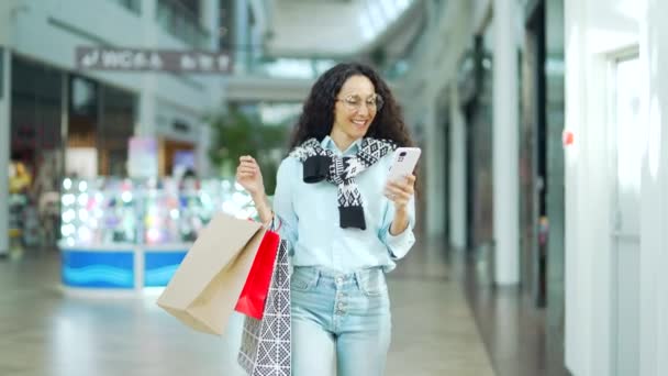 若い幸せな女性買い物客喜び顧客はショッピングセンターモールを歩き 携帯電話のブラウジングチャットスマートフォンを使用していますギフトカラーパッケージバッグで満足して店に行きます — ストック動画