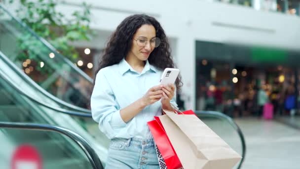 女性はショッピングセンターでスマートフォンを使用しています ショッピングバッグとアプリのスマート携帯電話を使用してモールブラウザで女性の買い物客を肖像画 幸せな女性購入スマイル満足顧客の店 — ストック動画