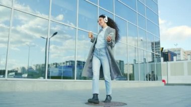 Neşeli genç İspanyol kadın modern bir şehir binasının arka sokaklarında dans ediyor ve elinde akıllı telefon ve bir fincan kahveyle kulaklıkla müzik dinliyor. Açık havada.