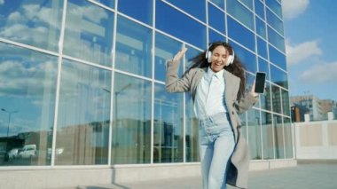 Neşeli genç İspanyol kadın şehir arka planında dans ederek neşe içinde yürüyor modern bir şehir binası ve kulaklıkla müzik dinliyor akıllı cep telefonuyla dışarıda neşeli, kaygısız bir yaşam tarzı var.