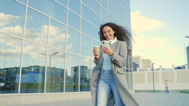 年轻漂亮的惊慌失措的女人走在城市街道的背景下 手拿着智能手机和咖啡杯享受生活 穿着灰色外套的女孩户外快乐生活的概念 — 图库视频影像