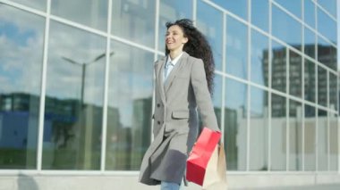 Mutlu, genç İspanyol kadın hediye kâğıdı torbalarıyla alışverişe çıkıyor. Arka planda gülümseyen, açık hava alışveriş merkezinde tatmin olmuş bir kadın. Modern bir alışveriş merkezi pazarında. Kara Cuma mutluluğu