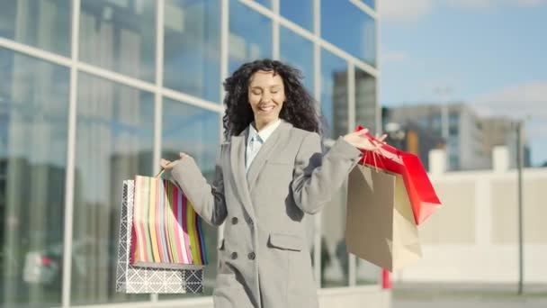 幸せな陽気な若いヒスパニック系の女性はギフトペーパーバッグで買い物をした後に行きます 屋外モールで満足した女性スマイリングは 現代のセンターセールスーパーマーケットを背景に歩いています ブラックフライデーの幸せ — ストック動画