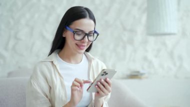 Gözlüklü, gülümseyen esmer kızın portresi akıllı telefonu tutar, salonda sosyal medyayı izler ve kaydırır. Açık tişörtlü genç bir kadın cep telefonunu evde kullanıyor ve ağda sohbet ediyor.