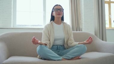Gözlüklü genç kadının portresi Lotus pozisyonunda oturma odasındaki kanepede otururken gözleri kapalı bir kadın evde meditasyon yogası yapıyor.