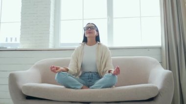 Gözlüklü genç kadının portresi Lotus pozisyonunda oturma odasındaki kanepede otururken gözleri kapalı bir kadın evde meditasyon yogası yapıyor.
