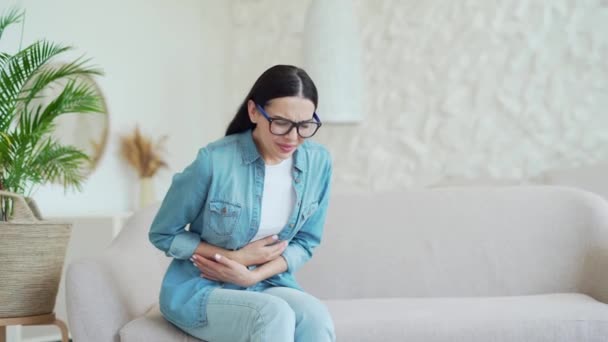 眼鏡の病気の暗い髪の女性は 胃の痛みがソファの上に座って苦しんで 腹を保持し 軽いリビングルームで胃炎や月経痛を感じています 痛みを伴う時代概念 — ストック動画