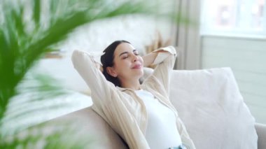 Koyu renk saçlı, güzel bir bayanın açık oturma odasında ellerini başının arkasına koyup rahat bir kanepeye uzanması. Gülümseyen barışçıl kız evde tek başına sakin vakit geçirmenin tadını çıkarıyor. Stres yok 