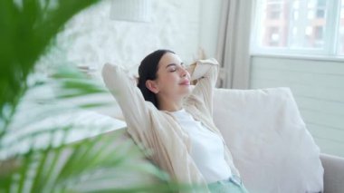 Koyu renk saçlı güzel bir bayan açık oturma odasında elleri başının arkasında rahat koltukta dinleniyor. Gülümseyen barışçıl kız evde tek başına sakin vakit geçirmenin tadını çıkarıyor. Stres yok.