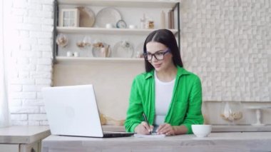 Koyu renk saçlı gözlüklü öğrencinin portresi, çevrimiçi konferans dinlerken ve evdeki laptop kamerasına bakarken uzaktan ders alıyor. Uzaklık eğitimi kavramı