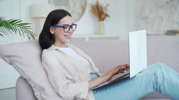 在客厅里躺在沙发上 用笔记本电脑对着眼镜的黑发女人微笑 有魅力的女性在网上工作或在家里上网 在线学习或在家工作 — 图库视频影像