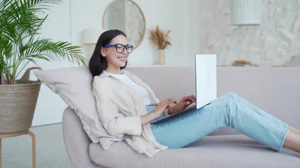 Gözlüklü Sırıtan Esmer Kadın Oturma Odasında Kanepede Yatarken Dizüstü Bilgisayar — Stok video