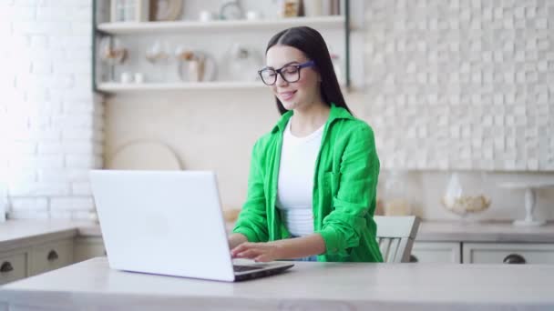 在光线室使用笔记本电脑 用眼镜对着深色头发微笑 有魅力的女人穿着绿色衬衫在网上工作或在家里浏览网页 在线远程学习或在家工作 — 图库视频影像