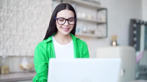 光の部屋でラップトップを使用して眼鏡でブルネットを微笑む 緑のシャツの中で魅力的な幸せな女性は オンライン仕事や家庭でウェブを閲覧を行う 屋内からのオンライン学習や仕事 — ストック動画