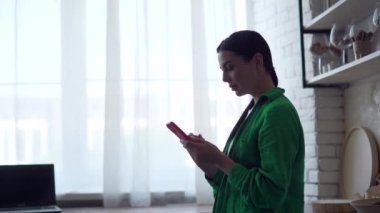 Genç bir kadının silueti akıllı telefonu tutuyor, e-postaları kontrol ediyor, sosyal medyayı izliyor, ev mutfağında kaydırıyor ve mesajlaşıyor. Kapalı alanda cep telefonu kullanan yeşil gömlekli çekici bir kadın.