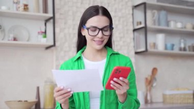Gözlüklü gülümseyen genç bir kadın faturaları ödüyor, ödeme işlemlerini açık oturma odasındaki telefonda online başvuru yaparak yapıyor. Finans yönetimi ve kolay maaş kavramı