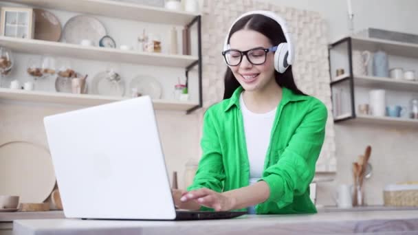 ノートパソコンで働く眼鏡の暗い髪の女性を笑顔と軽家庭のキッチンで音楽を聴く 自宅でオンライン仕事をするコンピュータ技術を使用して緑のシャツの若いフリーランサー — ストック動画