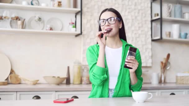 在家里的厨房里 一个深色头发 戴着眼镜的年轻女子在吃巧克力 这是她的画像 穿着绿色衬衫的漂亮女性独自咀嚼和享用黑巧克力 — 图库视频影像
