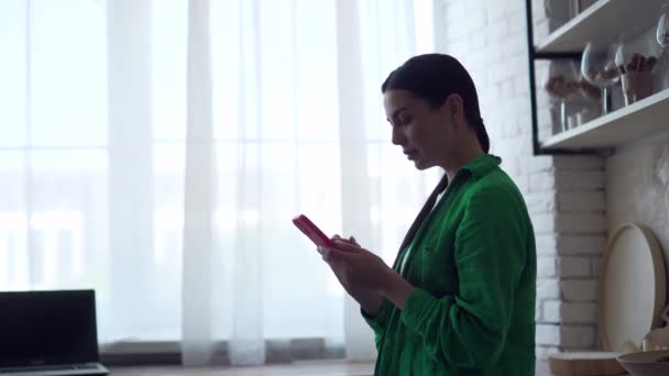 若い女性のシルエットは スマートフォンを保持し 電子メールをチェックし ソーシャルメディアを見て 国内のキッチンでスクロールしてテキストメッセージを送信します 携帯電話を屋内で使用して緑のシャツの魅力的な女性 — ストック動画