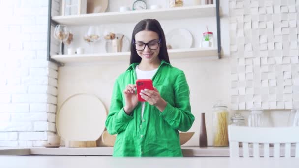 笑顔ブルネット女性女の子の肖像画で眼鏡をかけますスマートフォンスクロールを保持し 光の国内キッチンでソーシャルメディアを見て緑のシャツの若い女性を使用して携帯電話立って屋内 — ストック動画