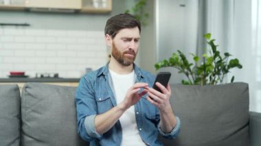 Bekar sakallı bir adam, flört uygulamasında kadın profillerini karıştırıyor ve oturma odasında akıllı telefondaki fotoğrafları beğeniyor ya da beğenmiyor. Sosyal platformda partner arayan ve eşleşme bekleyen genç bir adam.