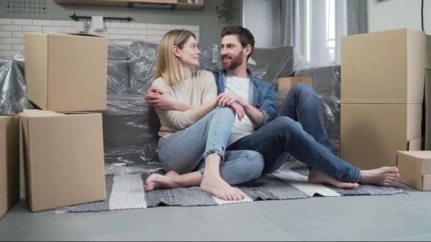 一对快乐的已婚夫妇坐在地板上 对搬进自己的房子或公寓 看着带着纸板箱的银行抵押贷款或房地产概念的相机 感到兴奋 — 图库视频影像
