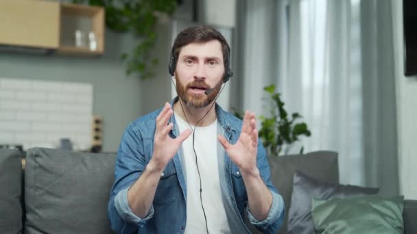 リビングルームでビデオ会議で話すビジネスコーチのウェブカメラビュー若い自信を持って髭の男は自宅でオンライン会議を持っている間 カメラを見て無線ヘッドセットを着用 — ストック動画