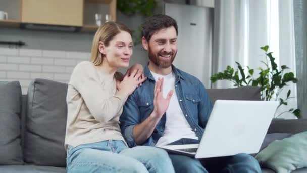 笑顔の若い家族のカップルは ラップトップカメラを見て 自宅でソファに座っているビデオ通話ウェブカメラオンライン会議で友人や家族に挨拶や話をする快適な会話のコンセプト — ストック動画