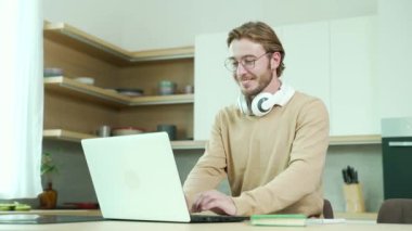 Dizüstü bilgisayarda çalışan mutlu genç serbest çalışan ev içi iş yerinde e-posta sörfü yapan gözlüklü, daktilo yazan ve evde otururken internette gezinen meşgul bir adam.