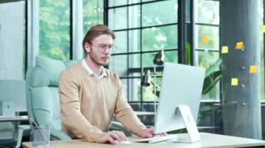 Çok çalışan, sırtı ağrıyan gözlüklü genç bir işadamı modern iş yerindeki bilgisayar başında çok çalıştıktan sonra iç mekandaki gerilimden muzdarip.