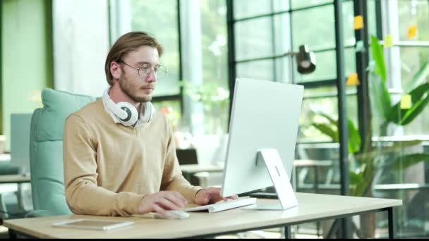 現代の職場でコンピュータに取り組んでいる彼の首にヘッドフォンで幸せな若いビジネスマン従業員デスクに座っている間 キーボード上でテキストメッセージやブラウジングを入力する忙しいオフィスワーカー起業家 — ストック動画
