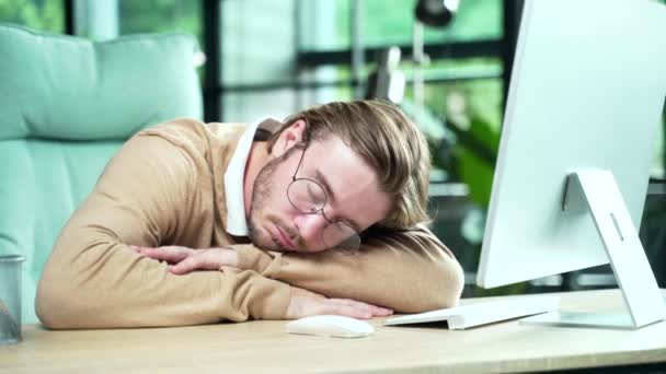仕事の時間に職場で寝そべっている眼鏡をかけた若いOlを疲れきったサラリーマンの肖像コンピュータの隣の机の上に寝転がっている男 — ストック動画